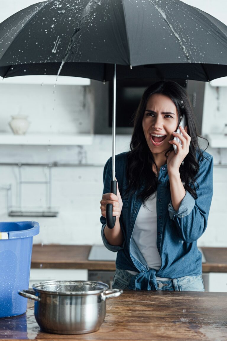 Stressed woman using umbrella during leak in kitchen and calling plumber